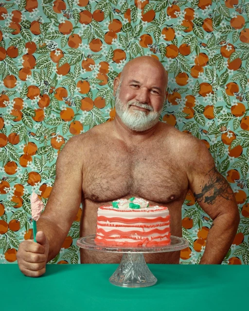 Man poses topless with a cake on a stand in front of a patterned wallpaper; he has a white beard and a tattoo on his arm.